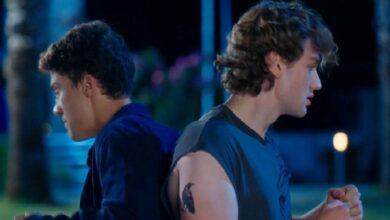 Two young men stand back‑to‑back outdoors at night, one in a blue shirt facing left and the other in a sleeveless gray shirt facing right, with a tattoo on the upper arm of the latter.
