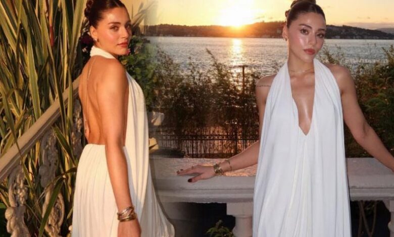 Two women in white halter dresses stand on a balcony at sunset, with a body of water in the background.