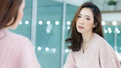 Asian woman wearing a pink knit sweater leans toward a mirror, gazing at her reflection in a bright room comprise of glass walls.