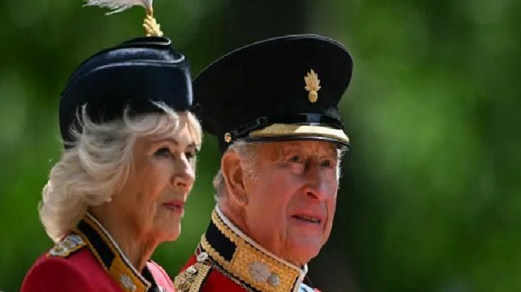 Elderly couple in red ceremonial military uniforms with gold braiding, outdoors with green background, side by side and looking ahead.