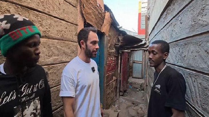 Three men stand in a narrow, sunlit alley between weathered wooden walls, engaged in conversation.
