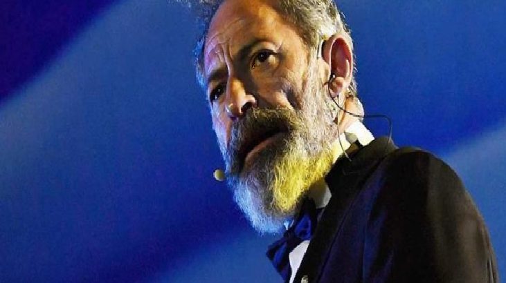 Bearded man in a suit on stage with a headset microphone, blue stage lighting behind him.