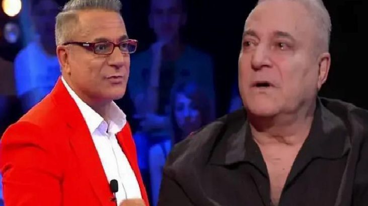 Two men on a TV talk-show set: one in a bright red blazer with glasses, the other in a black shirt, both wearing lapel mics amid an audience backdrop.