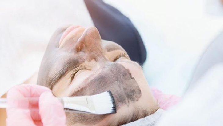 Person receives a mud facial as a brush applies a dark clay mask to their cheek and forehead with a gloved hand.
