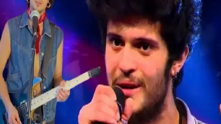 Male singer holding a microphone on stage during a live performance; guitarist in a denim vest plays in the background under blue stage lights.