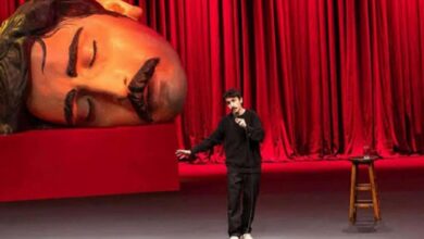 Comedian on stage with microphone, gesturing, beside a giant reclining statue head against red curtains.