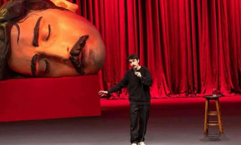 Comedian on stage with microphone, gesturing, beside a giant reclining statue head against red curtains.