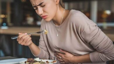 Woman grimacing and holding her stomach while eating a plate of food at a restaurant, indicating stomach pain or discomfort.