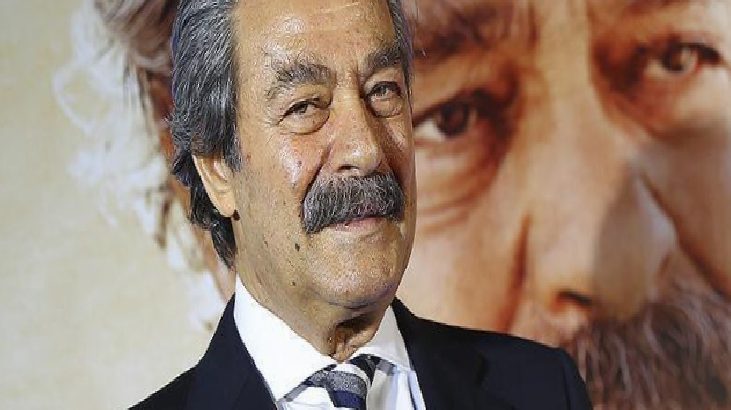 Older man with dark mustache in a suit and striped tie, posing in front of a large poster backdrop of another face.