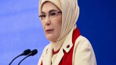 Woman wearing a beige hijab and red scarf speaks at a podium with two microphones against a blue background.