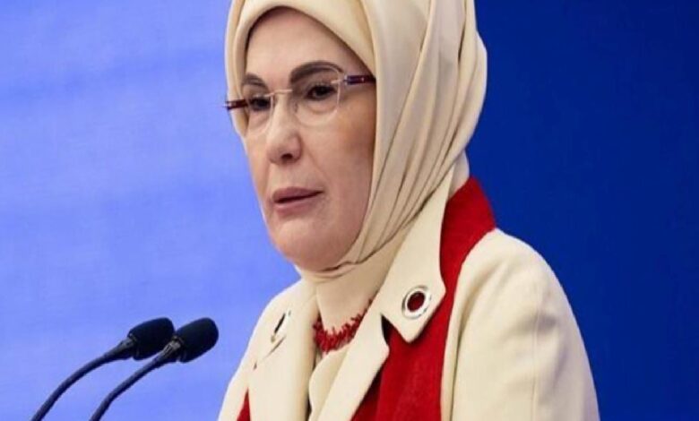 Woman wearing a beige hijab and red scarf speaks at a podium with two microphones against a blue background.