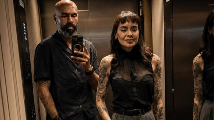 Bearded man in a black shirt takes a selfie in an elevator with a tattooed woman beside him in a sheer black top.