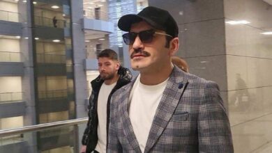 Man in a gray plaid blazer, white T-shirt, cap and sunglasses walking in a modern shopping mall.