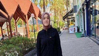 Woman wearing a light brown hijab and dark striped top stands on a sunlit outdoor shopping street behind storefronts and greenery.