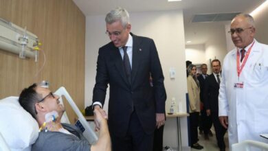 Smiling man in a suit shakes hands with a patient lying in a hospital bed; a doctor watches nearby.