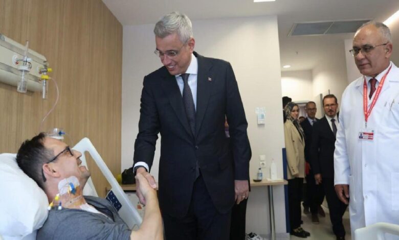 Smiling man in a suit shakes hands with a patient lying in a hospital bed; a doctor watches nearby.