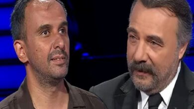 Two men stand close together on a TV set, one in a brown shirt and the other in a suit, under blue studio lighting.