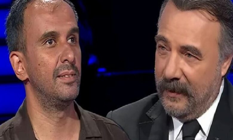 Two men stand close together on a TV set, one in a brown shirt and the other in a suit, under blue studio lighting.