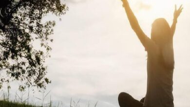 Person standing in a grassy field with arms raised toward the sun at sunset, signaling celebration or joy.