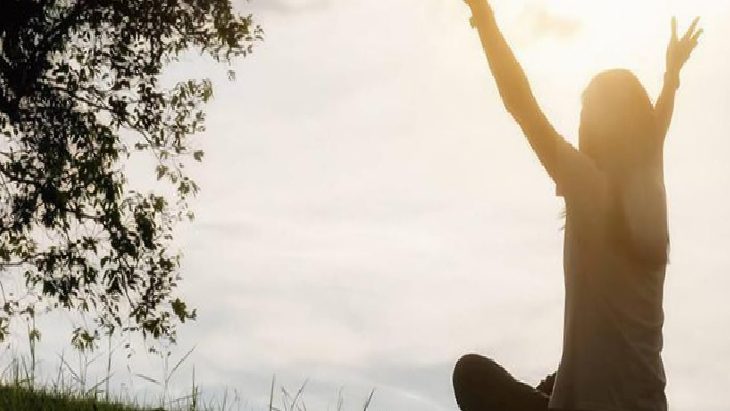 Person standing in a grassy field with arms raised toward the sun at sunset, signaling celebration or joy.