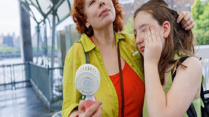 Two women outdoors; one woman comforts a tearful younger girl while holding a small portable fan.