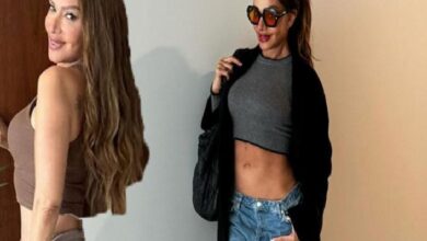 Two women posing together indoors: one with long brown hair in a brown tank top, the other in a gray crop top, blue jeans, a black coat, and sunglasses.