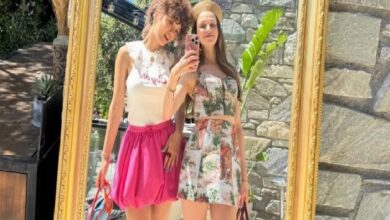 Two young women posing in front of a large ornate gold frame outdoors, wearing summer outfits (white top and pink skirt; floral dress).