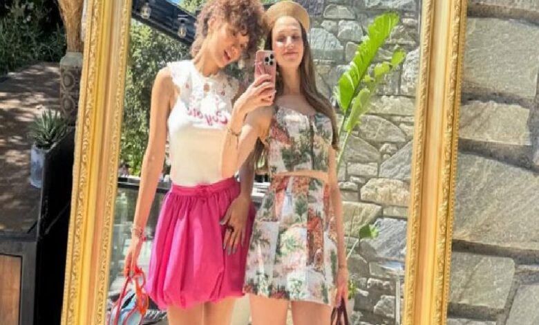 Two young women posing in front of a large ornate gold frame outdoors, wearing summer outfits (white top and pink skirt; floral dress).