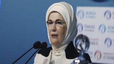 Woman in a white hijab and glasses speaking at a podium with multiple microphones, blue backdrop with logos behind her.