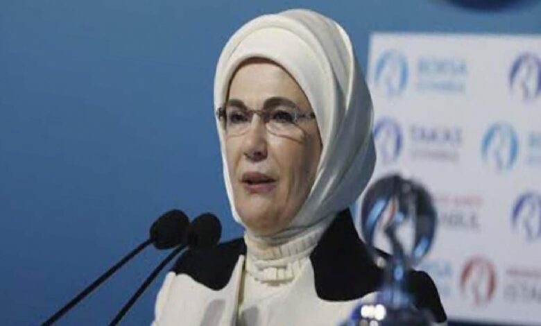 Woman in a white hijab and glasses speaking at a podium with multiple microphones, blue backdrop with logos behind her.