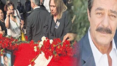 Mourners at a funeral with red flower arrangements and a framed photo; a photographer is in the background.