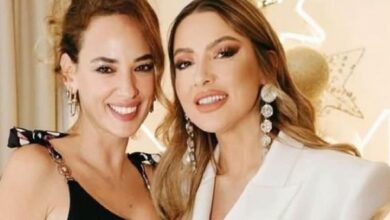 Two women posing and smiling at the camera at a formal event; one in a black top, the other in a white blazer with chandelier earrings.