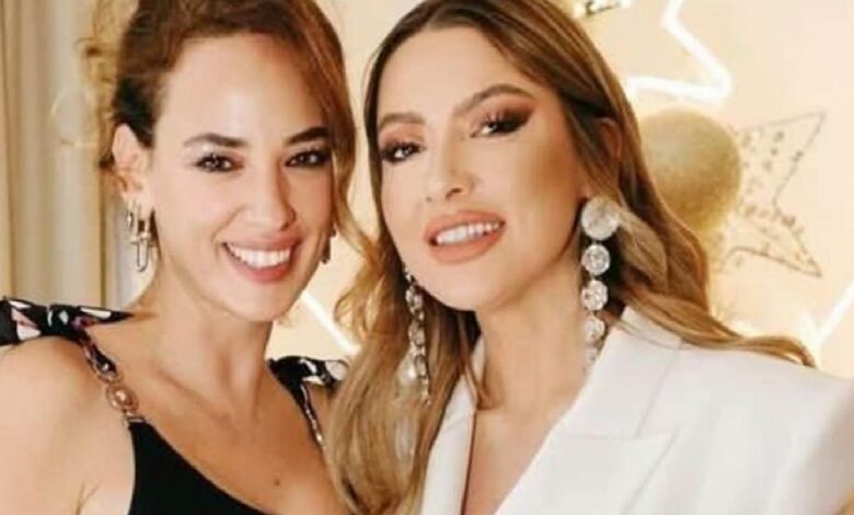 Two women posing and smiling at the camera at a formal event; one in a black top, the other in a white blazer with chandelier earrings.