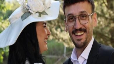 Smiling couple outdoors: woman wearing a large white hat with flowers and a white outfit, beside a man in a suit and glasses.