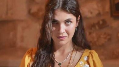Portrait of a woman with long dark hair, wearing a yellow dress and green jewelry, looking at the camera outdoors.