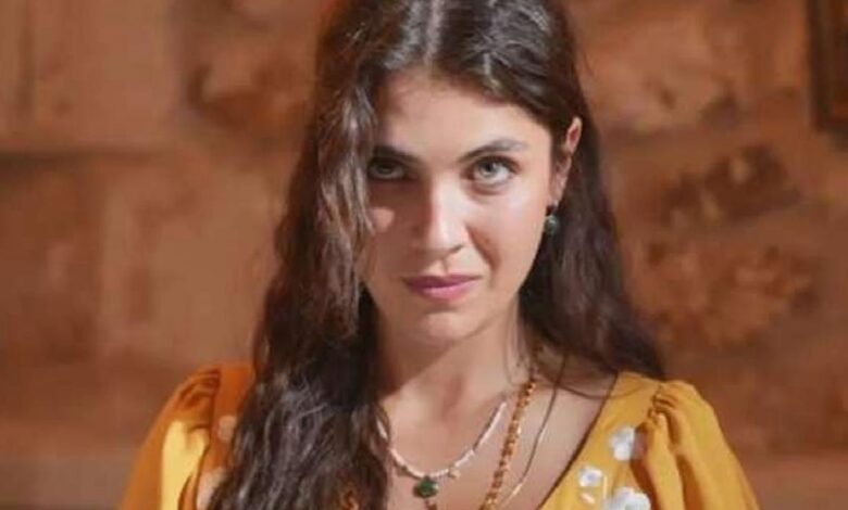 Portrait of a woman with long dark hair, wearing a yellow dress and green jewelry, looking at the camera outdoors.