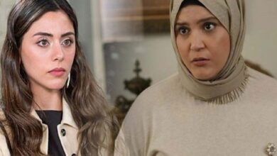 Two women stand indoors with serious expressions; the woman on the left has long brown hair and a light coat, the woman on the right wears a beige hijab and sweater.