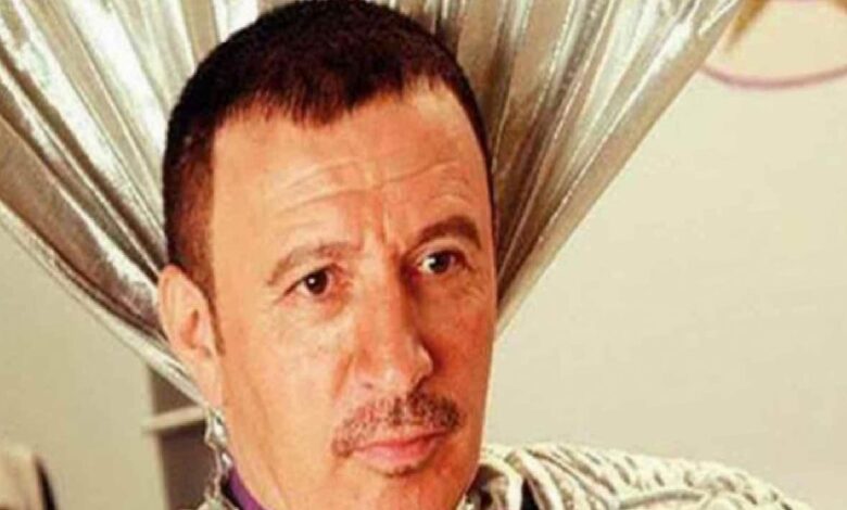 Man with short dark hair and mustache, looking to the right, indoors with beige background.