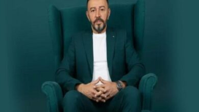Professional portrait: man in a dark teal suit seated in a matching high-back chair, hands clasped in his lap.