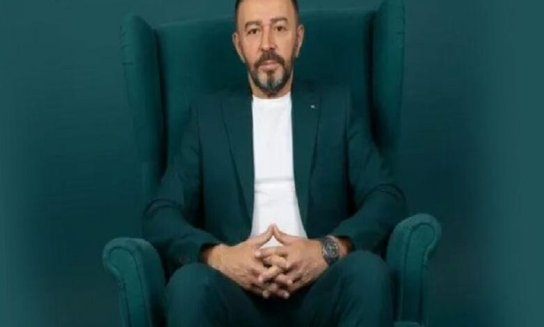 Professional portrait: man in a dark teal suit seated in a matching high-back chair, hands clasped in his lap.