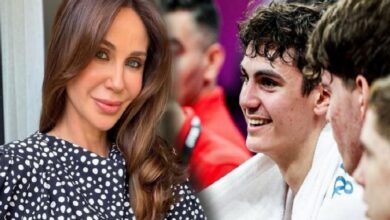 Woman with long brown hair in a polka-dot blouse standing beside a smiling young man in a white martial arts gi on a crowded sideline.