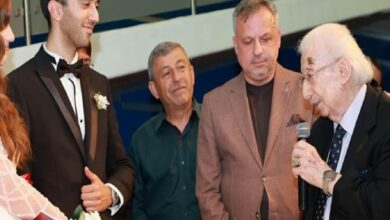 Groom in a black tux with a boutonniere stands with three men as an older man speaks into a microphone at a formal event/ceremony.