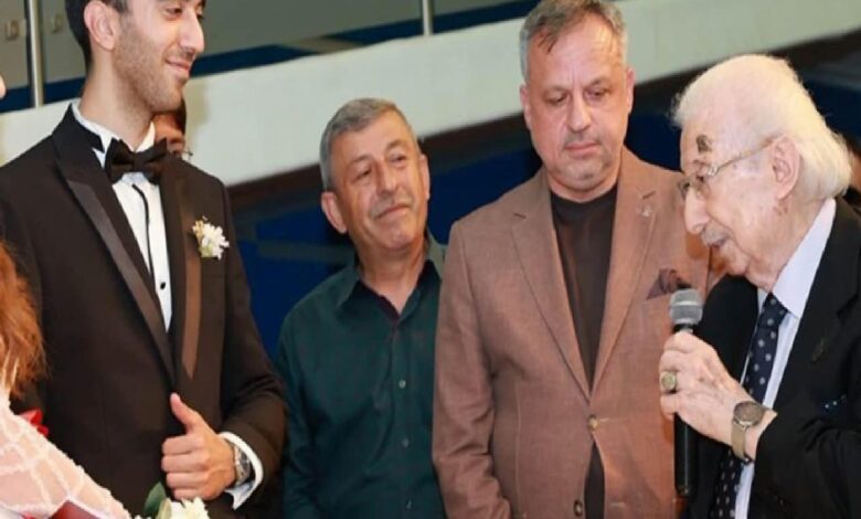 Groom in a black tux with a boutonniere stands with three men as an older man speaks into a microphone at a formal event/ceremony.
