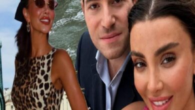 Three adults pose for a selfie outdoors by the water: a woman in a leopard print dress on the left, a man in a suit center, and a woman in pink on the right.