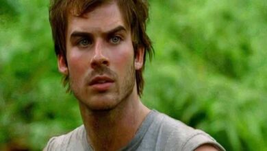 Young man with brown hair and stubble looks surprised in a green outdoor setting.