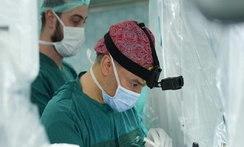Surgeon wearing a red patterned surgical cap, headlamp, and mask performing surgery with assistant nearby in a sterile operating room.