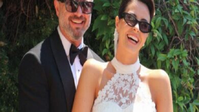 Smiling couple in formal attire at an outdoor wedding; man in a black tuxedo, woman in a lace white dress wearing sunglasses.