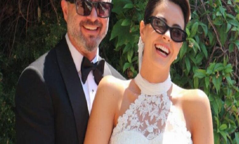 Smiling couple in formal attire at an outdoor wedding; man in a black tuxedo, woman in a lace white dress wearing sunglasses.