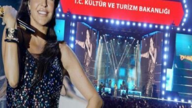Female singer on a bright concert stage, microphone in hand, wearing a sequined top, with large screens and a red banner in Turkish above.