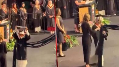 Graduate in cap and gown raises the hood on stage during a graduation ceremony, with other graduates and a speaker at a podium in the background.